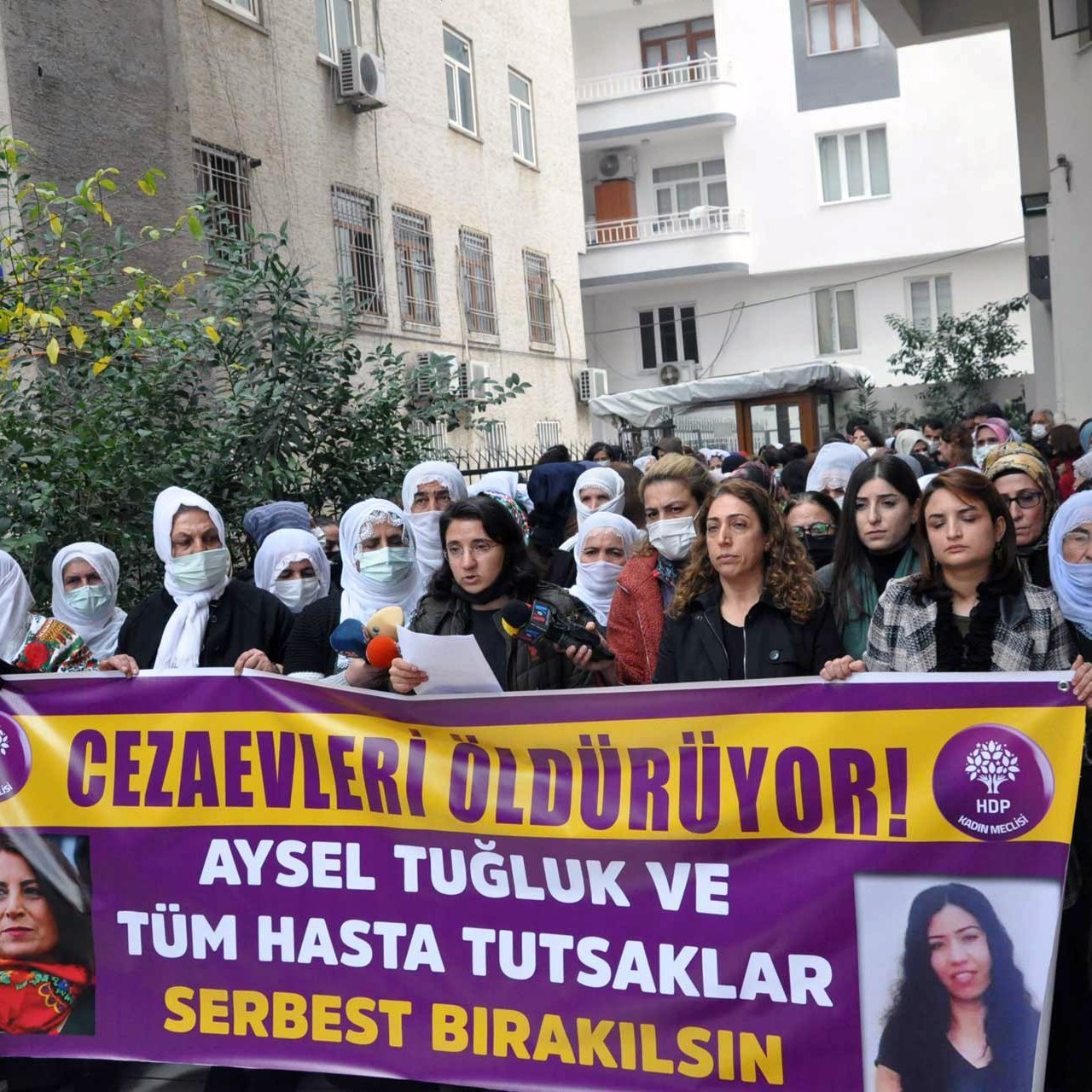 L'image montre un groupe de personnes rassemblées dans une rue, tenant des pancartes et des banderoles. Les femmes portent des foulards sur la tête et semblent soutenir une cause ou un mouvement. L'une des banderoles porte des slogans demandant la libération d'Aysel Tuğluk et des prisonniers malades. Le contexte semble être une manifestation pour les droits de l'homme et la justice. Des expressions de détermination et de solidarité sont visibles parmi les participants.