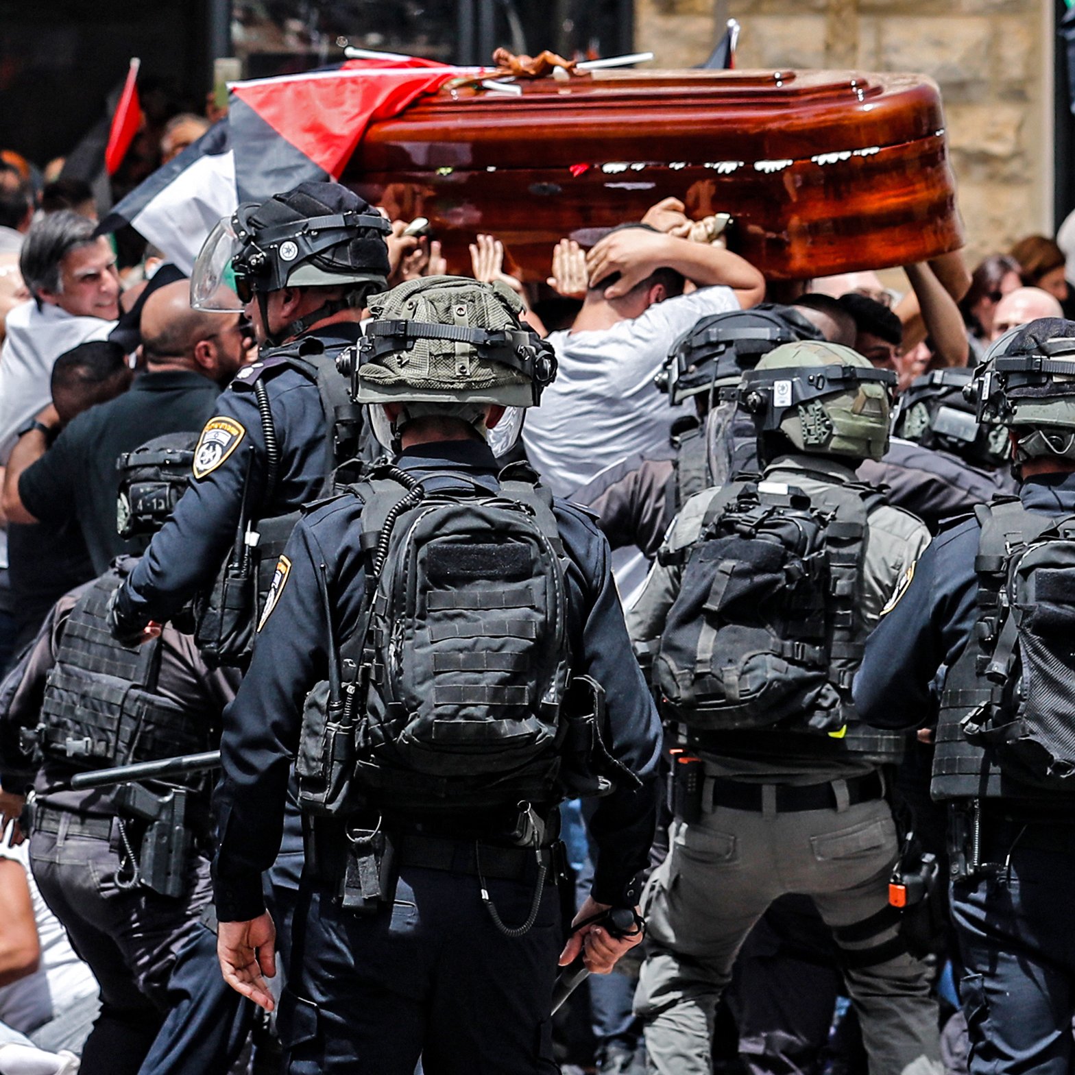 L'immagine rappresenta una scena di tensione durante una manifestazione. Ci sono persone che trasportano un barella, probabilmente durante un funerale o una commemorazione, mentre un gruppo di agenti di polizia in equipaggiamento anti-sommossa sta cercando di mantenere l'ordine. Molti manifestanti sono visibili, con bandiere che simboleggiano le loro rivendicazioni, e l'atmosfera appare carica di emozione e conflitto. La scena è caratterizzata da un forte senso di mobilitazione e protesta.