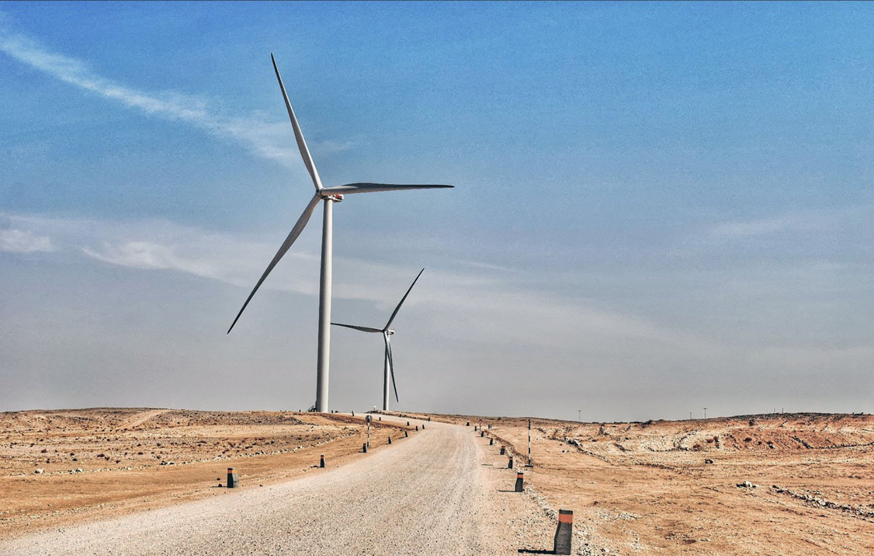 تظهر في هذه الصورة منظرًا طبيعيًا صحراويًا يمتد تحت سماء زرقاء فاتحة. يتواجد في الجزء الأمامي طريق رملي يتجه نحو الأفق، محاطًا بأعمدة صغيرة على جانبيه. في الخلفية، هناك عدة توربينات رياح ضخمة تدور ببطء، مما يُعطي انطباعًا حديثًا حول استخدام الطاقة المتجددة في هذا المناطق القاحلة. يبدو المكان هادئًا وواسعًا، مع تباين واضح بين الألوان الرمادية للأرض والألوان الزرقاء للسماء.