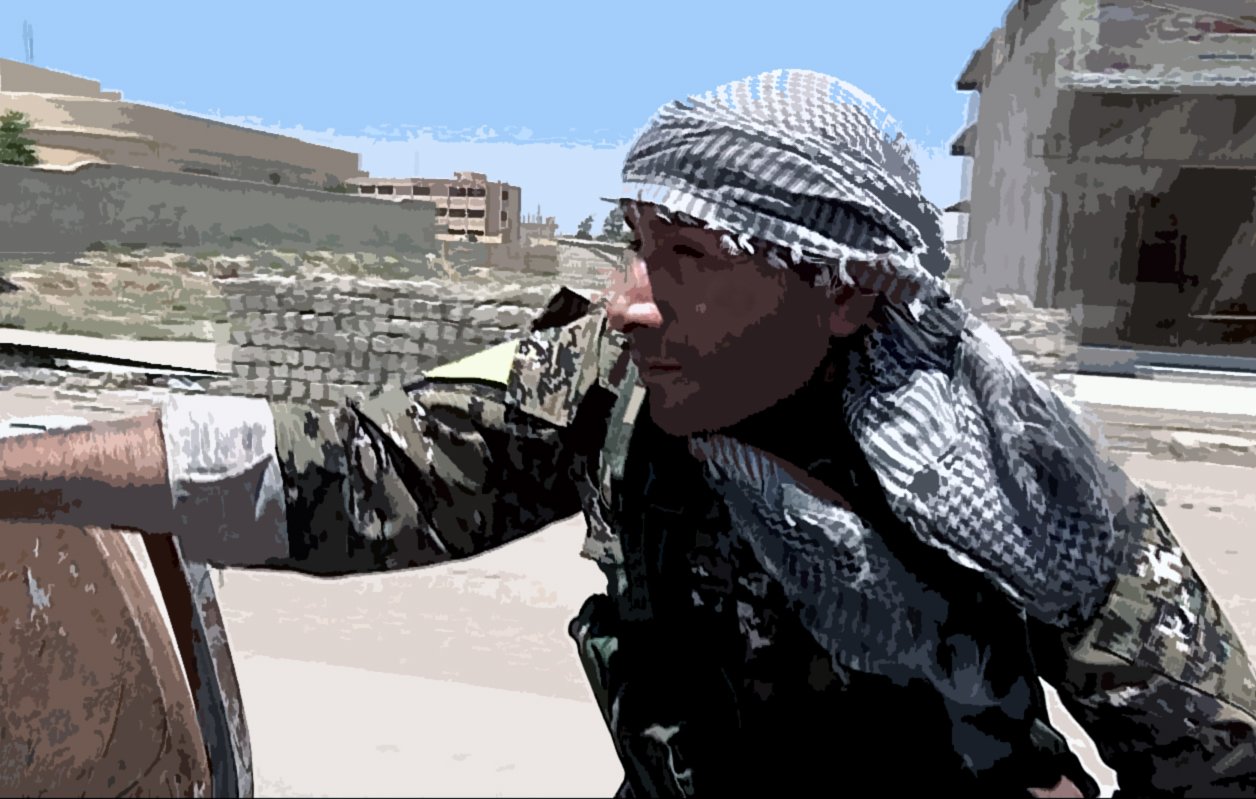 L'image montre un homme en tenue militaire, portant un foulard sur la tête. Il semble concentré et se trouve près d'un baril. En arrière-plan, on aperçoit des bâtiments et un paysage urbain. La scène évoque une ambiance de stress ou de tension, caractéristique des zones de conflit.