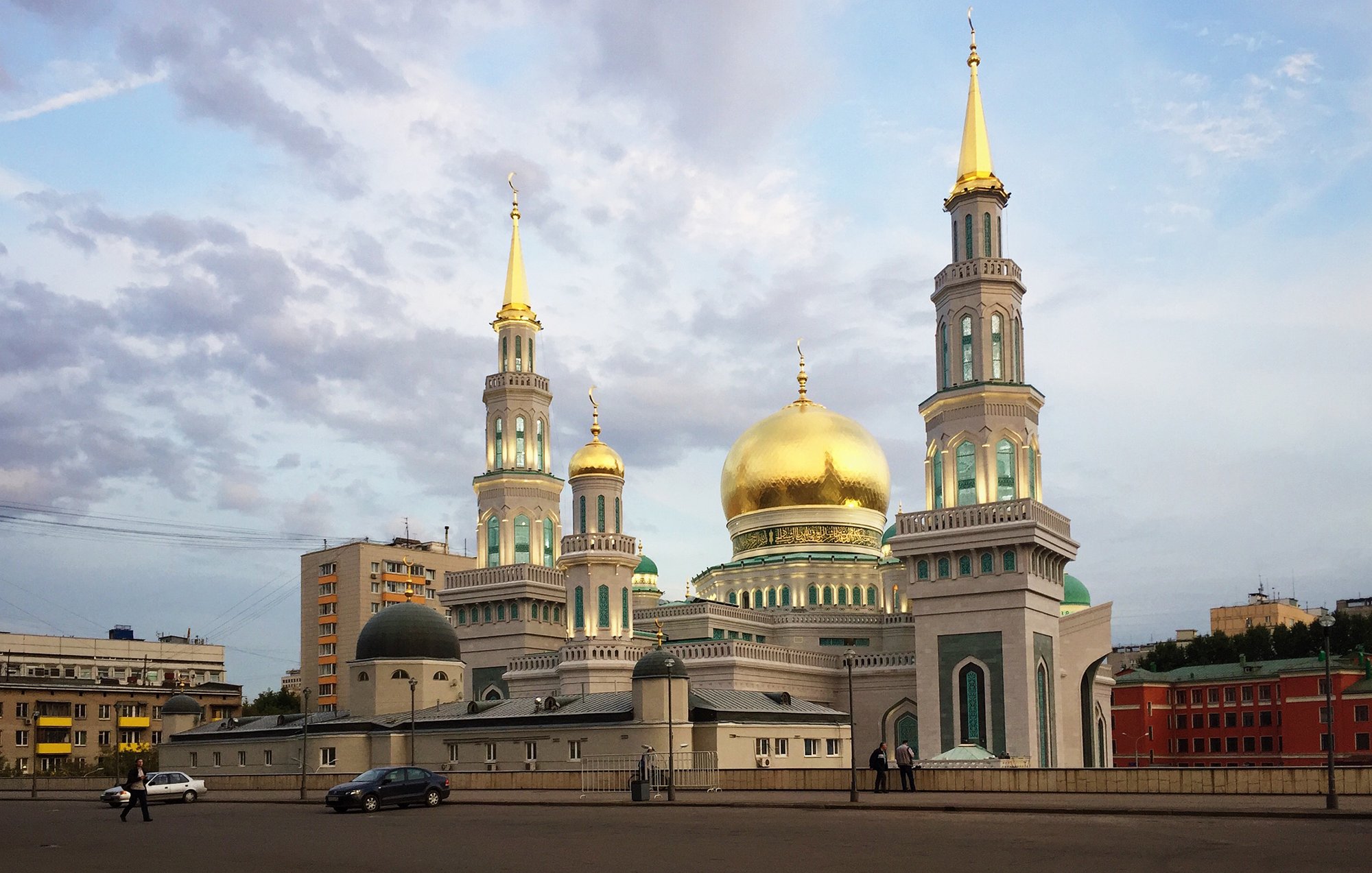 L'image montre une mosquée impressionnante avec des coupoles dorées et plusieurs minarets. Les structures sont ornées de détails architecturaux colorés, et le ciel en arrière-plan est légèrement nuageux, créant une ambiance tranquille. La mosquée se dresse majestueusement en milieu de ville, entourée d'immeubles modernes.