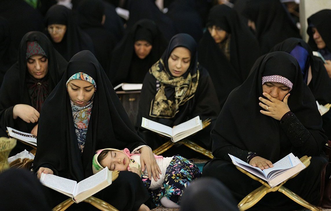 L'image montre un groupe de femmes, toutes vêtues de vêtements sombres, assises en train de lire des livres, vraisemblablement des copies du Coran. Certaines femmes sont profondément concentrées sur leur lecture, tandis qu'une d'elles tient un enfant qui semble s'endormir sur ses genoux. L'atmosphère est sérieuse et contemplative, indiquant un moment de prière ou de méditation collective. Les femmes portent des hijabs, ce qui est caractéristique de certaines traditions culturelles et religieuses. L'environnement semble être un espace communautaire, peut-être dans une mosquée ou un lieu de réunion.