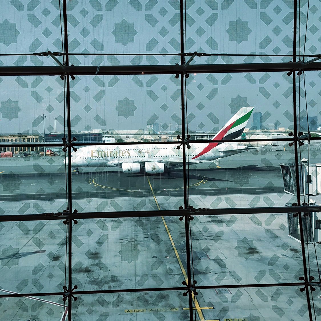 تظهر في الصورة منظر من داخل مطار، حيث يتبين في الخلفية طائرة تابعة لطيران الإمارات. يوجد زجاج مزخرف يزين النافذة، مما يضيف لمسة جمالية. يمكن رؤية بعض العربات والحقائب على أرضية المطار، مما يدل على حركة السفر والمغادرة. السماء تميل إلى الازرق وتظهر أجواء نهارية.