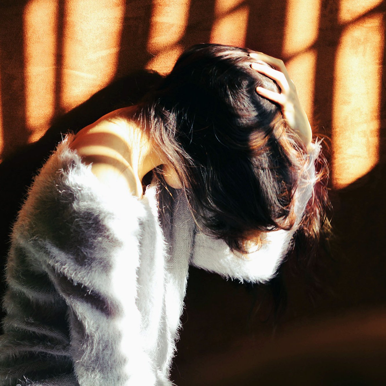 L'image montre une femme assise, penchée en avant et se tenant la tête avec ses mains. Sa posture suggère un état de réflexion ou de malaise. La lumière du soleil filtre à travers des barreaux, créant des ombres graphiques sur elle et sur le mur derrière. Elle porte un vêtement doux et pelucheux, ce qui contraste avec l'intensité de la lumière. L'ambiance générale de l'image évoque une certaine introspection ou une atmosphère mélancolique.