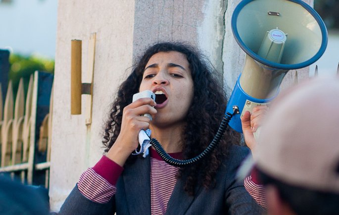 L'image montre une personne debout, utilisant un mégaphone pour s'adresser à un groupe. Elle semble engagée et passionnée, portant un costume avec une chemise à rayures. Son geste et son expression faciale indiquent qu'elle est en train de faire un discours, probablement lors d'une manifestation ou d'un rassemblement. En arrière-plan, des personnes écoutent attentivement, suggérant qu'elle est en train de discuter d'un sujet important. Le cadre général dénote une atmosphère de mobilisation ou d'activisme.