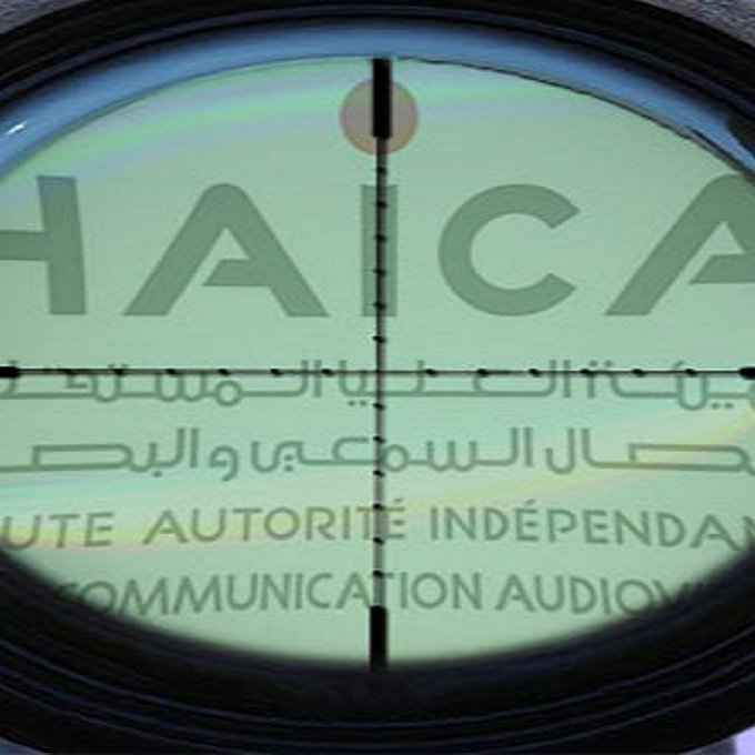 L'image montre un réticule de visée, typique d'une lunette de tir, qui se superpose à un fond avec le mot "HAICA" visible. Ce mot est associé à une autorité indépendante en matière de communication auditive, peut-être en rapport avec la régulation des médias. Le contraste entre l'optique et le texte suggère un thème de surveillance ou de contrôle. Le fond semble comporter divers éléments, peut-être liés à des événements ou des institutions médiatiques.