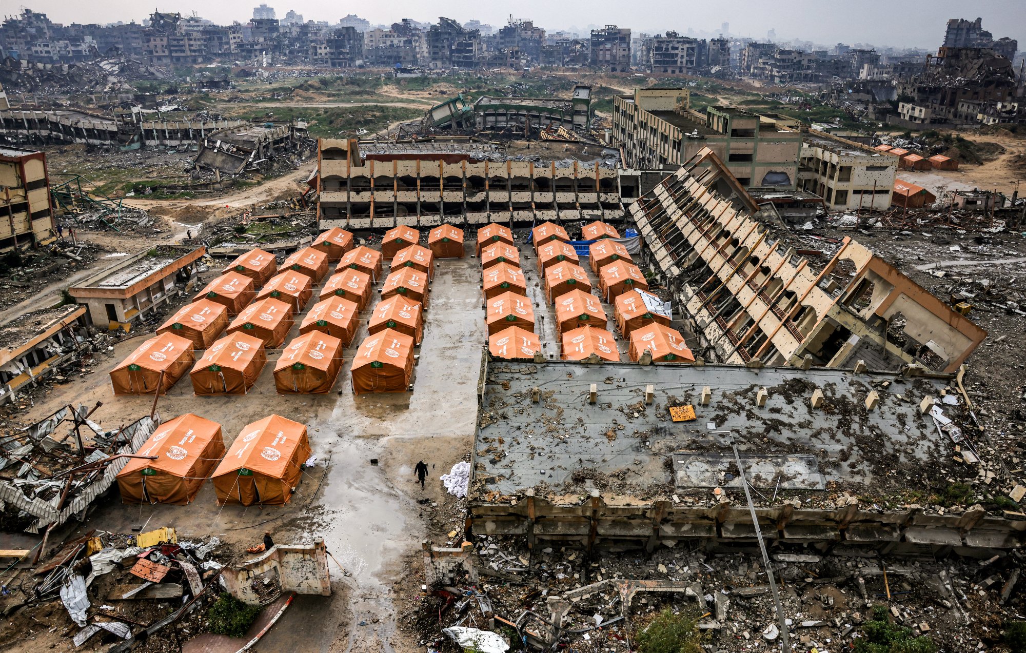L'immagine mostra un'area devastata, con edifici in rovina sullo sfondo e tende arancioni disposte ordinatamente in primo piano. Il terreno sembra sporco e disordinato, evidenziando una situazione di emergenza o di sfollamento.