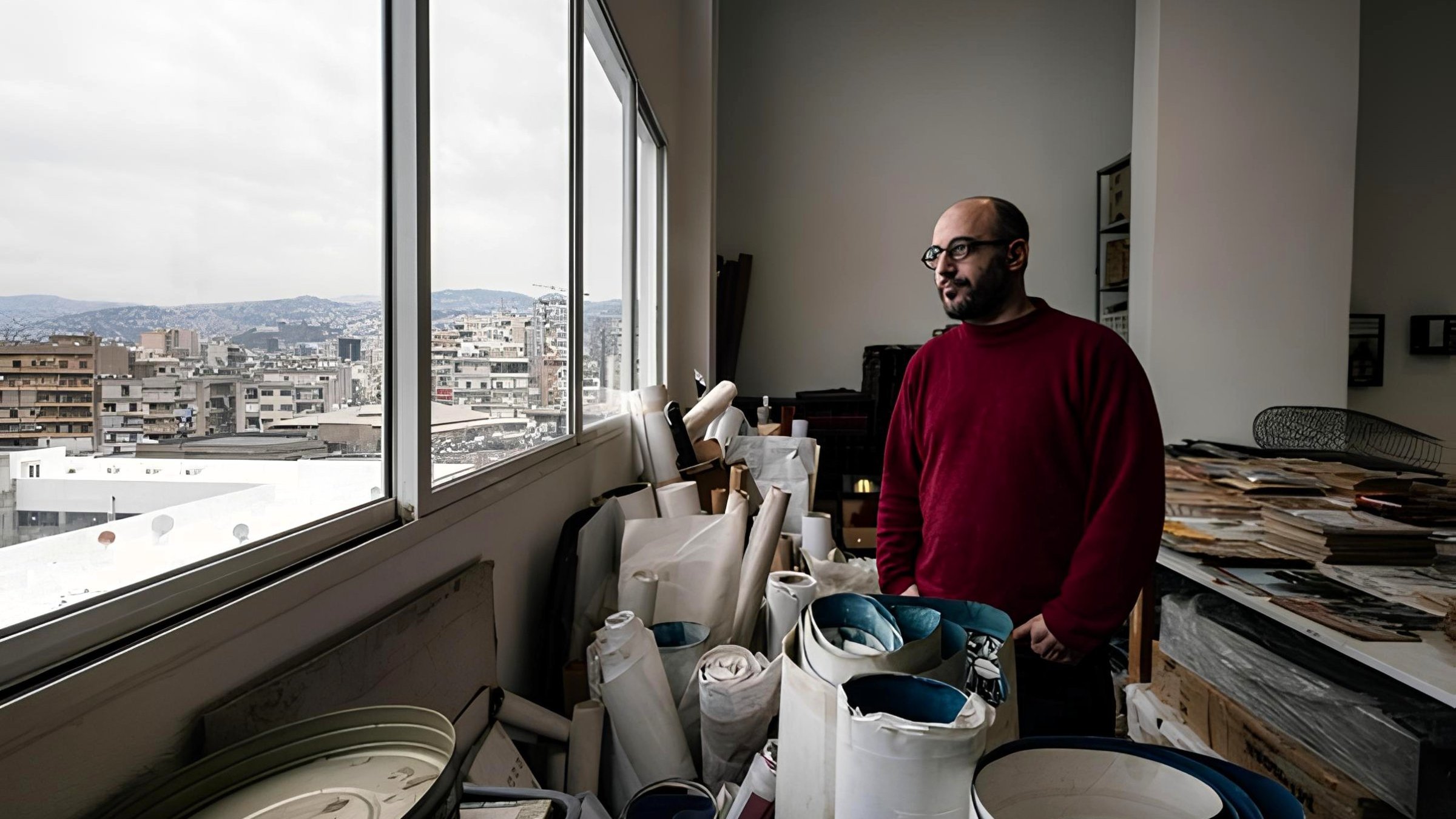 Uomo in maglione rosso guarda fuori da una finestra, circondato da rotoli di carta e materiali.