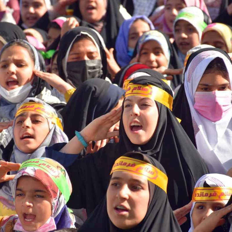 L'image montre un grand groupe de jeunes filles, portant des hijabs et des bandeaux avec des inscriptions en arabe. Elles semblent exprimer de l'enthousiasme, chantant ou criant, et leurs visages sont pleins d'énergie. L'arrière-plan indique un rassemblement, probablement lié à un événement spécial ou une manifestation. Les couleurs des vêtements et des bandeaux apportent une atmosphère vibrante à la scène.