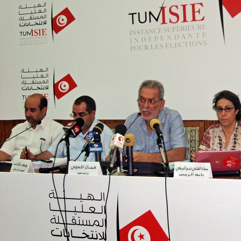 L'image montre une conférence de presse avec plusieurs personnes assises à une table. Un homme au centre semble parler, tandis que les autres personnes à ses côtés écoutent ou prennent des notes. Il y a des microphones sur la table et un arrière-plan avec des logos et du texte en arabe et en français, probablement lié à une institution électorale. L'environnement semble formel et consacré aux affaires publiques.