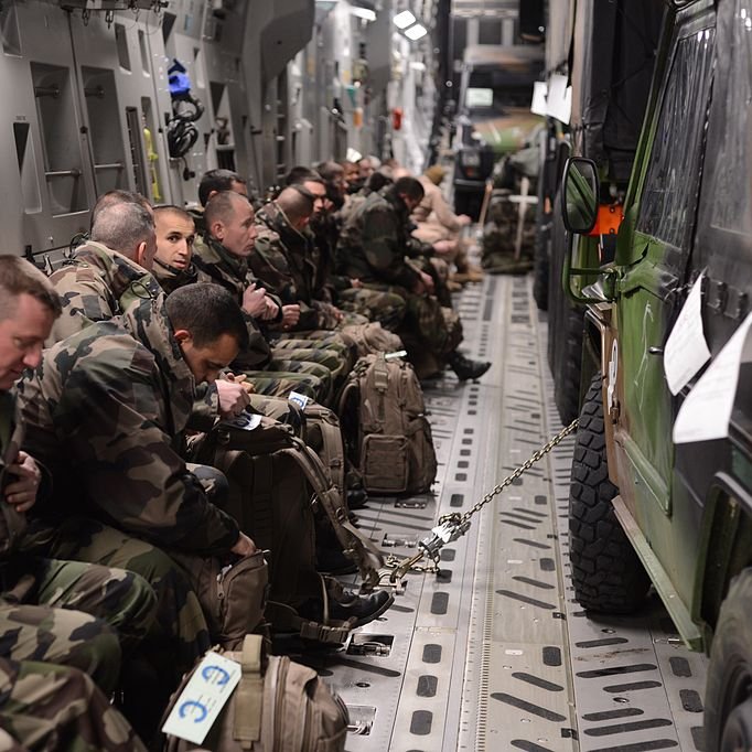در این تصویر، گروهی از سربازان در داخل یک هواپیما یا وسیله نقلیه نظامی نشسته‌اند. سربازان در لباس‌های camouflage (رنگ‌های نظامی) دیده می‌شوند و به سمت جلو نشسته‌اند، در حالی که هر کدام یک کوله‌پشتی دارند. فضای داخلی وسیله نقلیه به نظر مدرن می‌رسد و تجهیزات نظامی در اطراف آن قرار دارد. برخی از سربازان به نظر می‌رسد در حال آماده‌باش بوده و توجه‌شان به جلو معطوف است.