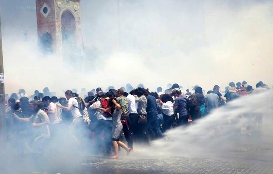 L'image montre une scène de manifestation où un grand groupe de personnes semble être en mouvement, probablement en train d'échapper à des gaz lacrymogènes ou à une intervention des forces de l'ordre. Le brouillard blanc, causé par la fumée ou les sprays, rend l'atmosphère tendue et chaotique. En arrière-plan, une structure architecturale est visible, mais elle est à peine discernable à cause de la fumée. Les manifestants semblent rassemblés et stressés, certains essayant de se protéger ou de se diriger vers une zone plus sûre.
