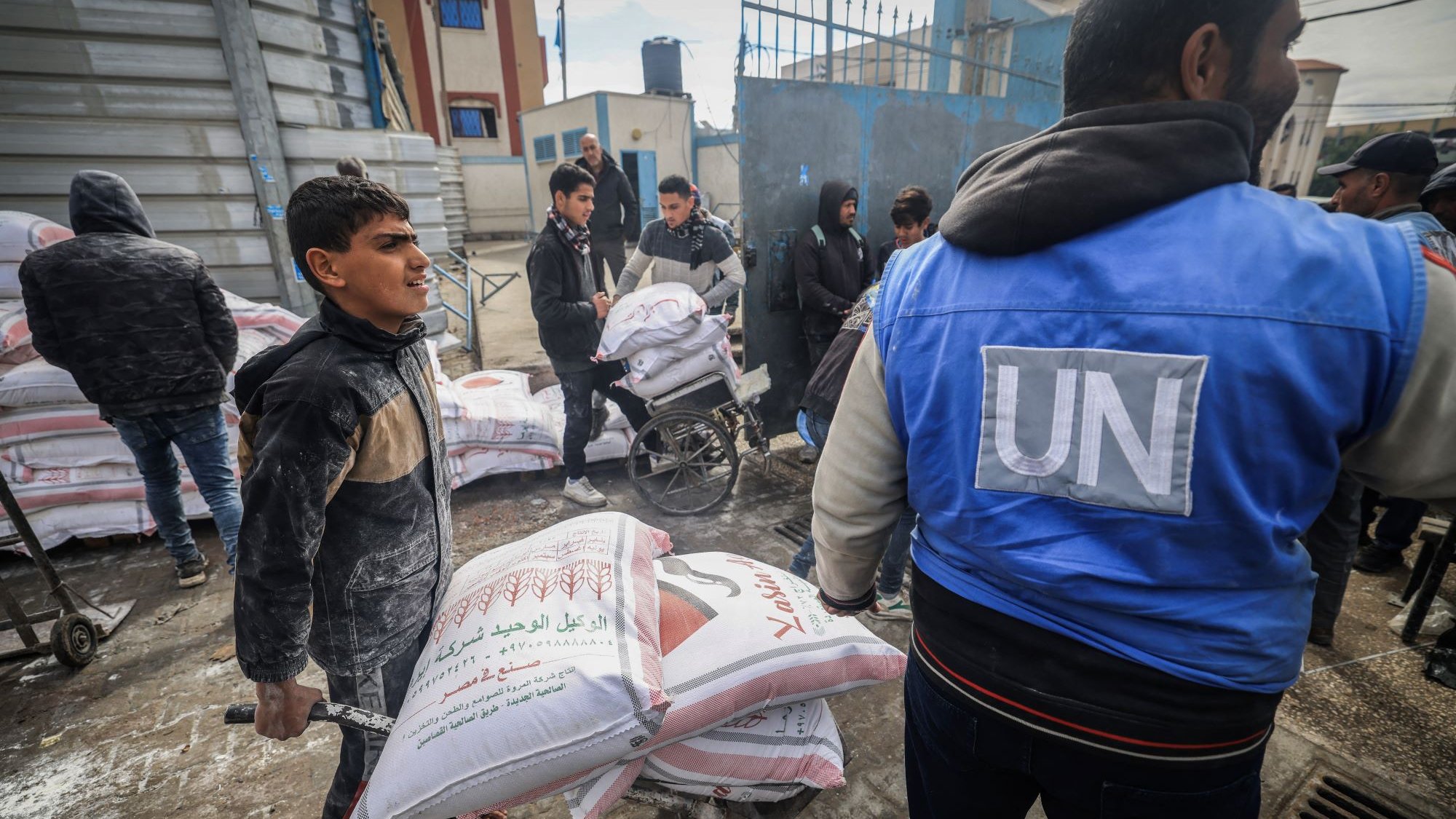 L'immagine mostra una scena di aiuto umanitario. Alcuni ragazzi e adulti sono impegnati nel trasporto di sacchi, probabilmente di cibo o materiali di prima necessità, all'interno di una zona urbana. Un ragazzo sta spingendo un carrello con sacchi, mentre altre persone, alcuni dei quali indossano giubbotti con il logo delle Nazioni Unite, sono visibili in background e stanno trasportando ulteriori carichi. La scena comunica un senso di collaborazione e supporto nella distribuzione di aiuti umanitari.