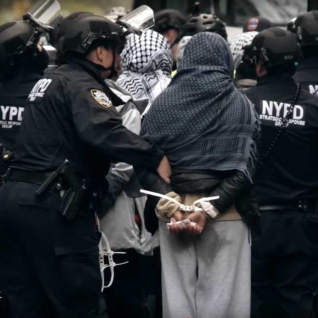 L'image montre un groupe de policiers en uniforme noir entourant une personne arrêtée. Cette personne est de dos, les mains attachées derrière. Les policiers portent des casques et des protections, et il y a une atmosphère tendue, suggérant une manifestation ou une intervention policière.