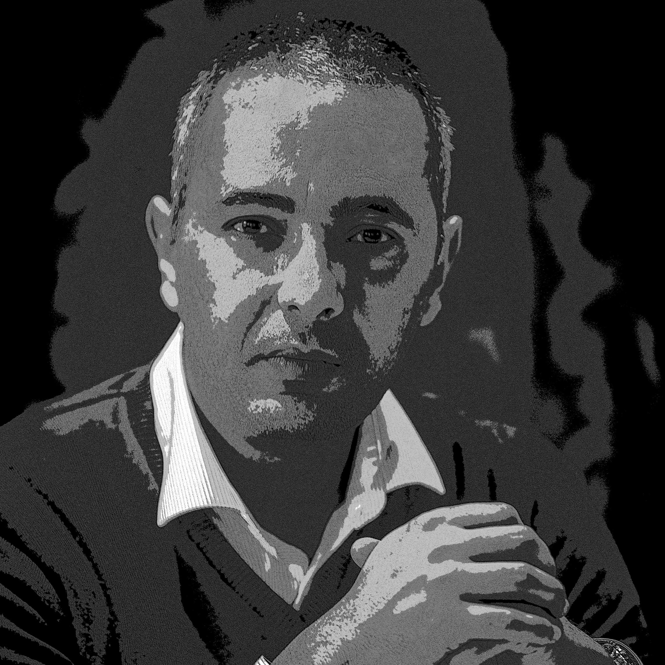 L'image montre un homme avec une expression réfléchie. Il a les cheveux courts et porte un pull. Ses mains sont croisées devant lui. Le fond est sombre, ce qui met en avant son visage. L'éclairage accentue ses traits et donne une atmosphère sérieuse à la photo.