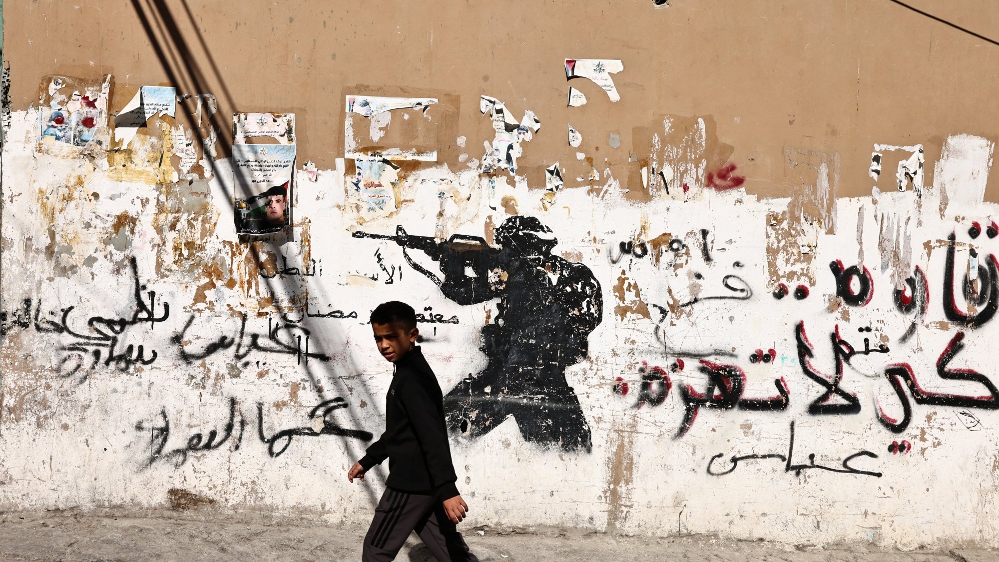 A boy walks past a wall with a soldier graffiti and Arabic text, contrasting street life.