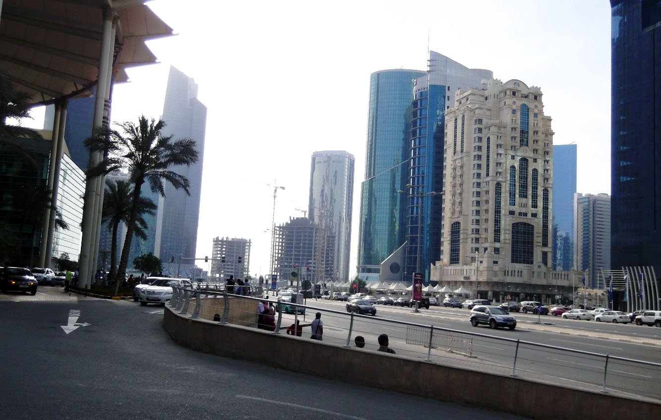 The image depicts a busy city street featuring a mix of modern skyscrapers and architectural styles. On the left, there are palm trees and a wide road leading into the city. Various cars are seen driving, indicating active traffic. The tall buildings on either side showcase glass and steel facades, reflecting light from the environment. The overall atmosphere suggests a bustling urban area with a contemporary vibe. The sky appears bright, contributing to the lively scene.