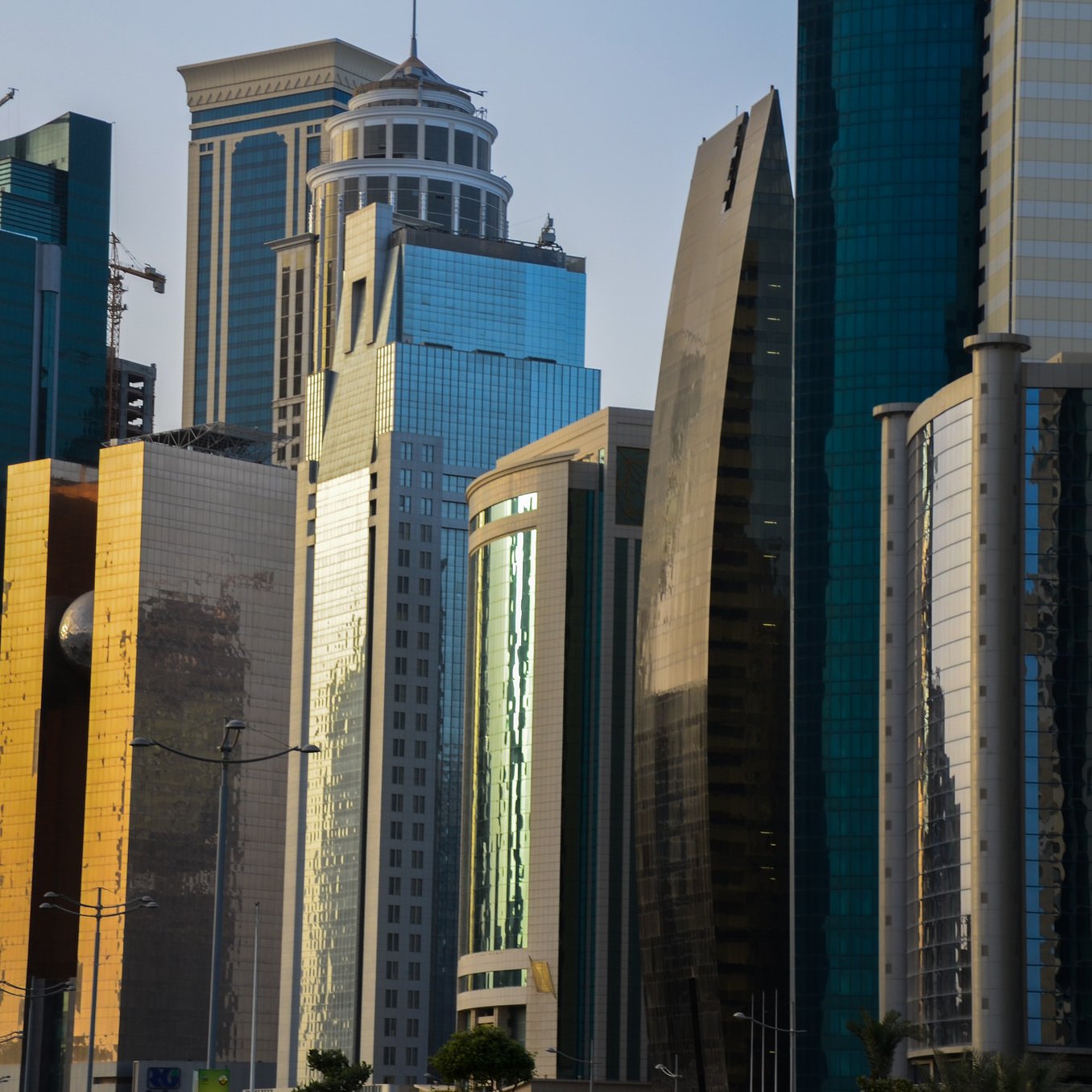 L'image montre une vue urbaine moderne avec plusieurs gratte-ciels aux façades brillantes et réfléchissantes. Les bâtiments sont de différentes formes et tailles, avec des matériaux en verre et en métal qui scintillent sous la lumière. On peut remarquer un mélange d'architectures contemporaines, certaines avec des courbes élégantes et d'autres plus angulaires. Le paysage urbain reflète un environnement dynamique et en pleine croissance, typique des grandes villes modernes.