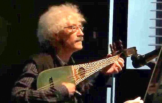 L'image montre un homme âgé avec des cheveux bouclés et gris, jouant d'un instrument de musique à cordes. Il porte des lunettes et une chemise à motifs, ainsi qu'un gilet. On peut voir une ambiance de concert ou de performance, avec un éclairage qui met en valeur l'artiste et son instrument, qui ressemble à un luth.