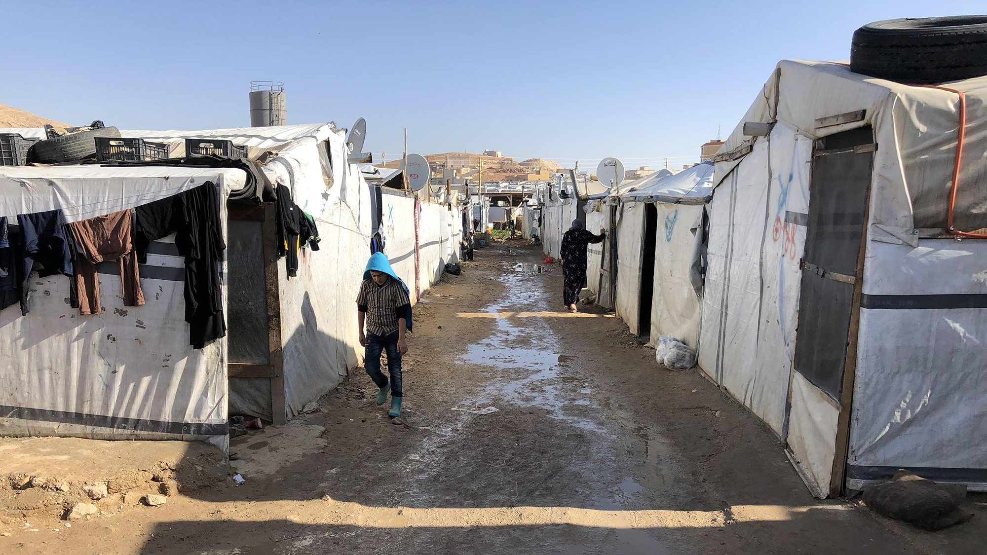 این تصویر نمایی از یک کمپ پناهندگان را نشان می‌دهد. در اینجا، چادرهای سفید و مشکی به صورت ردیفی در دو طرف یک مسیر خاکی قرار دارند. زمین به نظر می‌رسد کمی مرطوب و گل‌آلود است. چندین قطعه لباس بر روی چادرها آویزان شده‌اند و شخصی در حال حرکت در مسیر دیده می‌شود. در پس‌زمینه، ساختمان‌هایی در بالای تپه‌ها قابل مشاهده هستند. این منظره فضایی ساده و دلگیر را تداعی می‌کند.