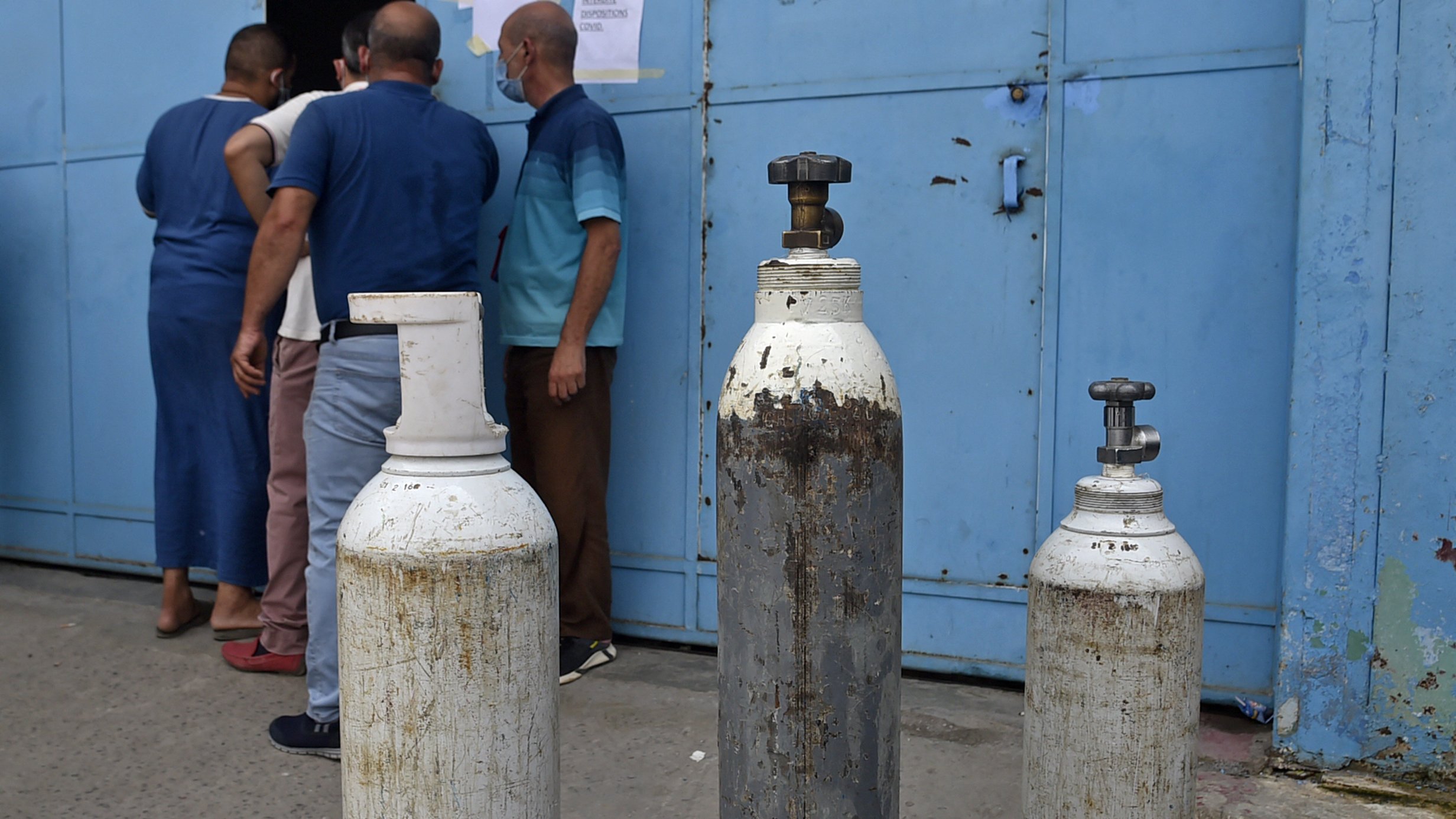 تظهر الصورة مجموعة من أسطوانات الغاز التي تُستخدم عادةً في المستشفيات أو للأغراض الصناعية. توجد ثلاث أسطوانات بأحجام متفاوتة، وتبدو واحدة منها قديمة ومُهملة. في الخلفية، يمكن رؤية بعض الأشخاص يقفون بالقرب من باب مبلوطة باللون الأزرق، مما يشير إلى موقع ربما يكون مركزًا لتوزيع الغاز أو خدمة طبية. تبدو الأجواء حيّة مع تواجد الأشخاص، مما يُبرز أهمية تلك الأسطوانات في الحياة اليومية.