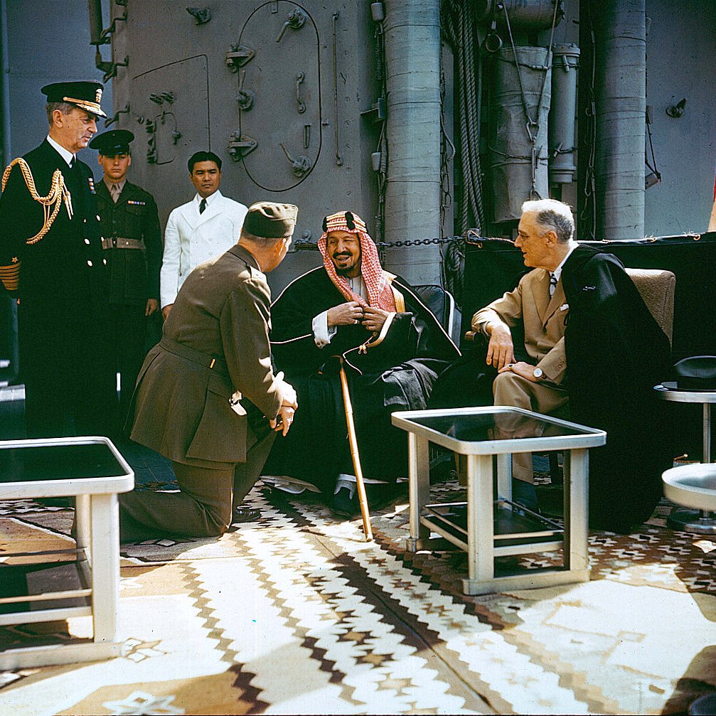 L'image montre un groupe de personnalités en discussion sur un navire. Au centre, un homme en tenue traditionnelle semble rire ou sourire, assis avec une canne. À sa gauche, un homme en habit sombre, tandis qu'un autre, en uniforme militaire, est à genoux devant eux. En arrière-plan, d'autres personnes observent la scène, dont un officier en uniforme. Le décor est richement décoré avec des tapis et des éléments nautiques.