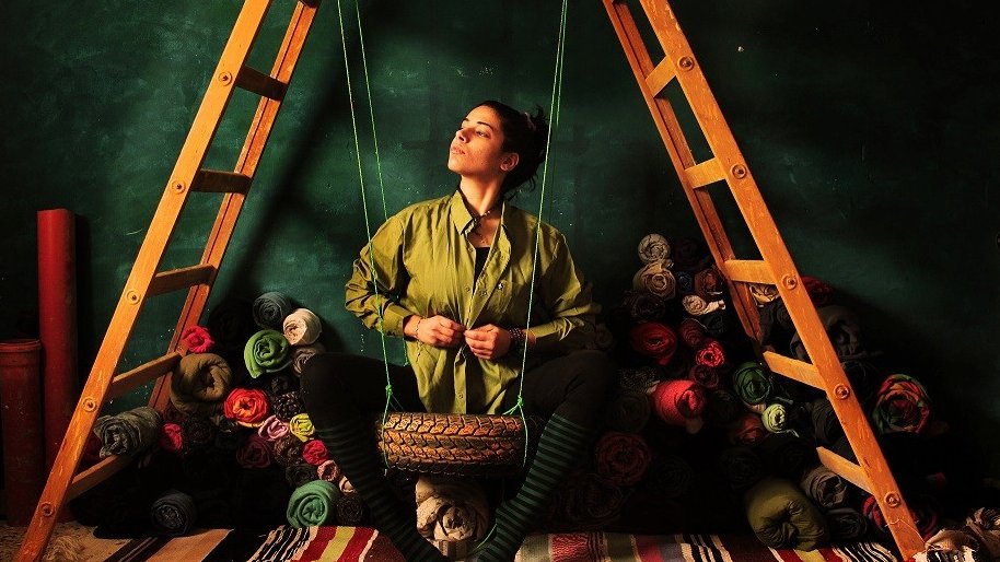 L'image représente une femme assise sur un hamac en forme de coussin, entre deux échelles en bois. Elle porte une chemise verte et semble pensive, le regard tourné vers l'horizon. En arrière-plan, des rouleaux de tissus colorés se trouvent empilés, ajoutant une touche de texture et de couleur à la scène. Le décor, avec des murs de couleur sombre, met en valeur le contraste avec les vêtements et les tissus, créant une ambiance artistique et contemplative.