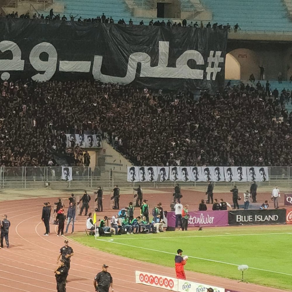 A large crowd at a stadium with a black banner and fans cheering passionately.