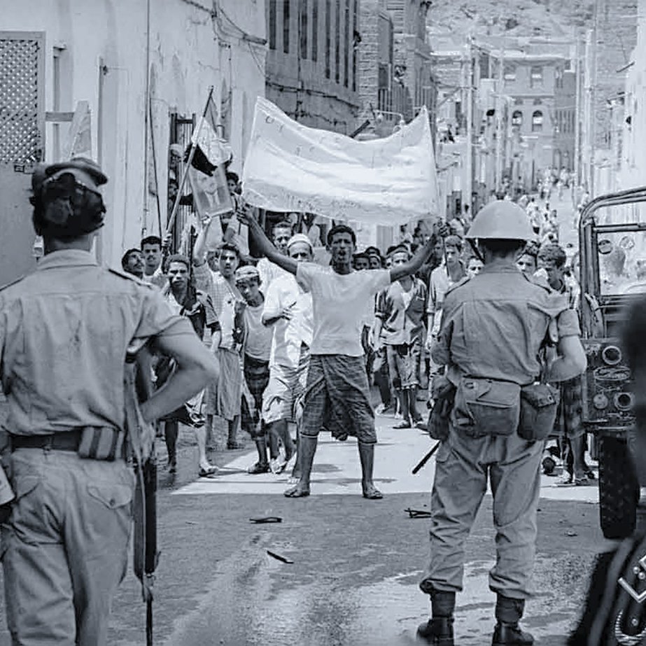 تظهر الصورة مجموعة من الأشخاص في شارع ضيق، يبدو أنهم في خضم حدث أو تظاهرة. في المقدمة، هناك رجل يرفع يديه حاملاً لافتة، بينما يتواجد أفراد من قوات الأمن بالقرب منه. يمكن رؤية الأطفال والبالغين في الخلفية، بعضهم يبدو مهتماً بالحدث. الملابس والإعداد تعكس فترة زمنية معينة، كما يظهر في التفاصيل المعمارية للمنازل. المشهد يحتوي على أجواء من الحماس والتوتر.