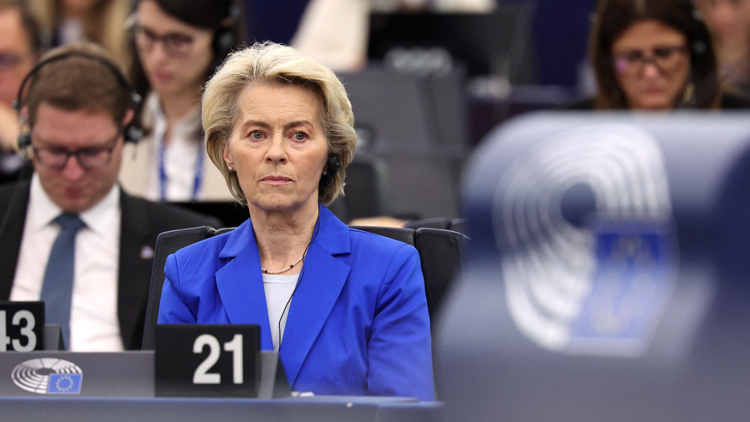 Nell'immagine si vede una donna seduta in un ambiente di tipo istituzionale, probabilmente durante una sessione del Parlamento Europeo. Indossa un blazer blu e ha i capelli biondi. Sembra concentrata e seria, mentre intorno a lei ci sono altre persone che ascoltano attentamente, alcune con cuffie. L'ambientazione è moderna, con posti a sedere e schermi visibili sullo sfondo.