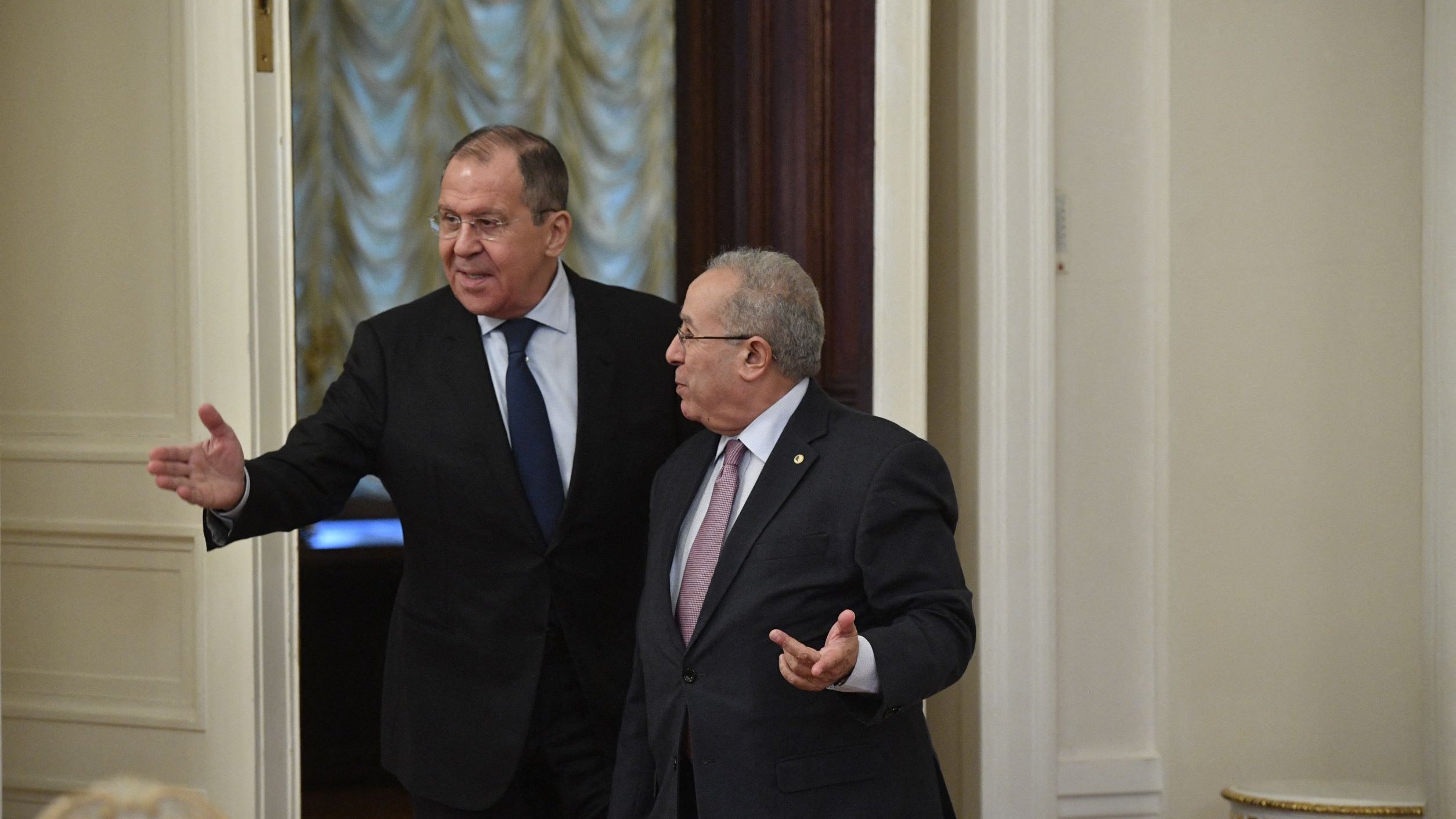 الصورة تظهر شخصين في غرفة داخلية ذات تصميم كلاسيكي. يبدو أن أحدهم يتحدث ويشير بيده، بينما الآخر يستمع. الإضاءة في الغرفة دافئة، وهناك ستائر ونوافذ خلفهم. الملابس الرسمية تشير إلى أن اللقاء قد يكون ذو طابع رسمي أو دبلوماسي.