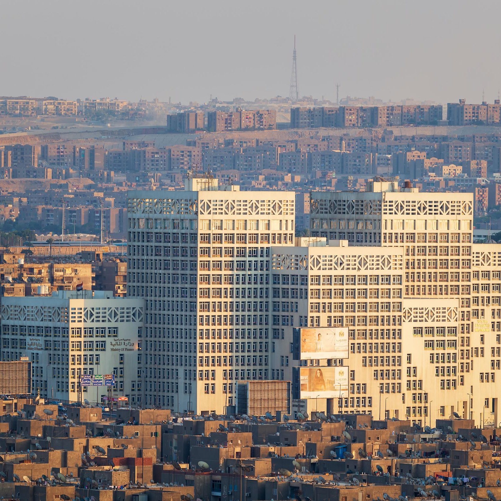 The image depicts a cityscape featuring a cluster of modern buildings in the foreground, characterized by their distinct architectural design with large glass windows. These buildings stand out against a backdrop of densely packed smaller structures in varying shades of brown. The scene conveys a contrast between contemporary architecture and older, more traditional urban forms. The setting appears to be at a distance, possibly showing a skyline during early morning or late afternoon when the light casts warm tones on the buildings. The surrounding area is marked by hilly terrain and a sprawling development, illustrating a bustling urban environment.