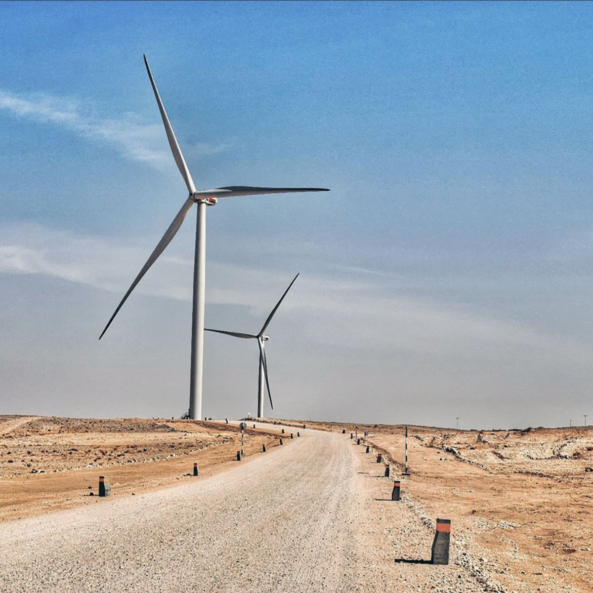 L'image montre un paysage désertique avec une route en gravier qui s'étend vers l'horizon. De chaque côté de la route, on aperçoit plusieurs éoliennes qui se dressent majestueusement contre un ciel bleu. Le terrain est aride, et les éoliennes symbolisent l'énergie renouvelable dans cet environnement désertique. Les colonnes le long de la route semblent délimiter le chemin, ajoutant une touche de structure à ce paysage naturel et technologique.