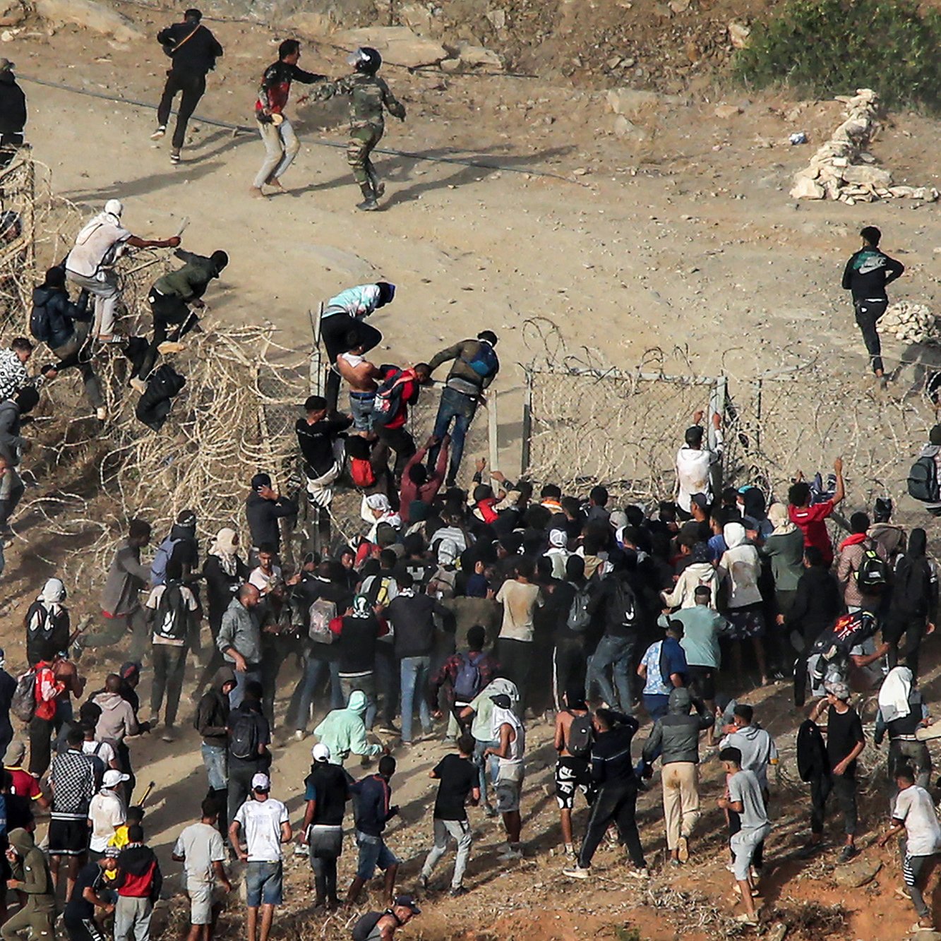 L'image montre un groupe de personnes tentant de franchir une barrière ou une clôture. On peut voir des individus escalader la clôture, tandis que d'autres semblent se rassembler en dessous. Il y a également des agents de sécurité présents à proximité, semblant surveiller la situation. L'environnement est celui d'une zone de passage, avec des éléments de terrain et des groupes de personnes manifestement en mouvement. L'atmosphère semble tendue, suggérant une situation d'urgence ou de conflit.