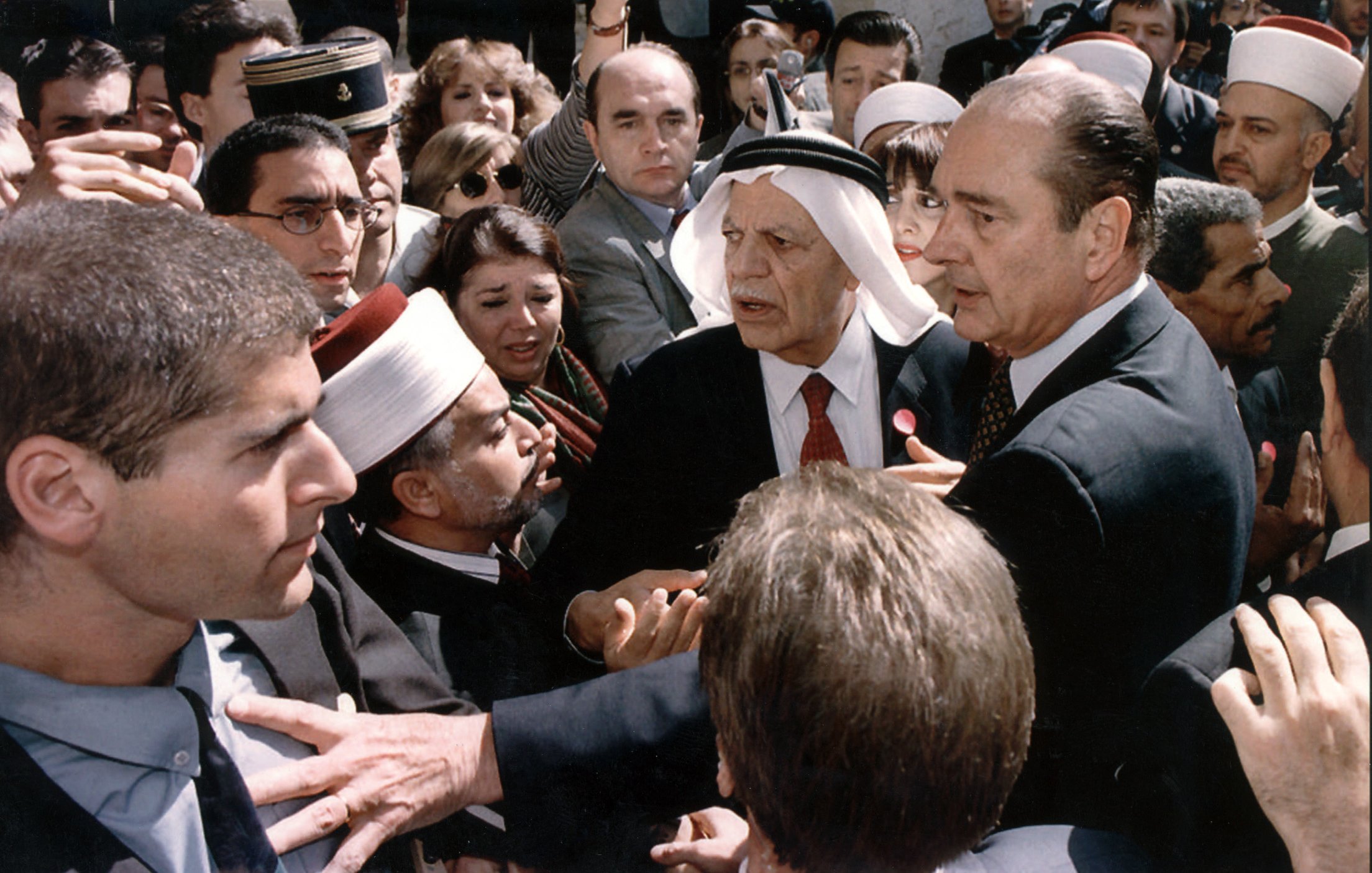 L'image montre une scène chargée d'émotion et de tension. On y voit plusieurs personnes rassemblées autour d'une figure centrale, visiblement un homme en costume. D'autres personnes, portant des vêtements traditionnels et des turbans, semblent interagir avec lui. L'atmosphère est dynamique, avec des visages concentrés et des gestes animés, ce qui suggère une discussion ou un événement important. Le mélange de styles vestimentaires indique une diversité culturelle parmi les personnes présentes, ce qui pourrait refléter un moment significatif sur le plan social ou politique.