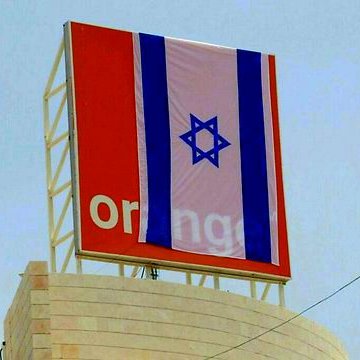 L'image montre un panneau affichant le logo de la marque "Orange", sur lequel est drapé un grand drapeau israélien. Le fond semble être un ciel clair, et le drapeau présente des bandes horizontales en bleu et blanc avec l'étoile de David au centre. Le tout donne une impression d'affichage public dans un environnement urbain.