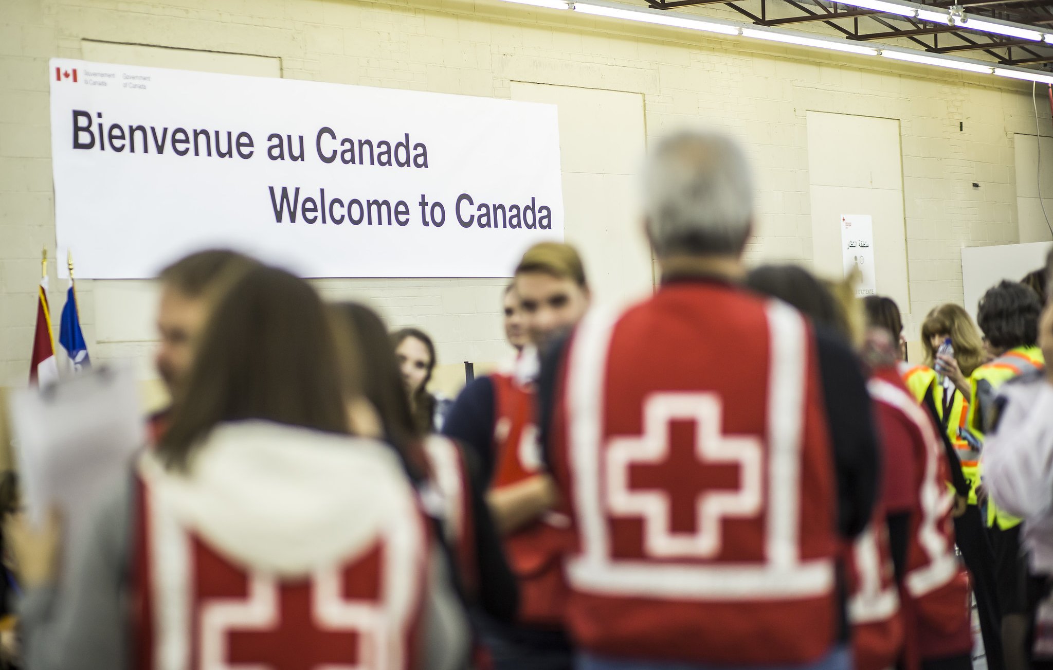L'image montre un intérieur où un groupe de personnes se rassemble. Au fond, il y a une grande affiche indiquant "Bienvenue au Canada" en français et en anglais. Plusieurs personnes portent des vestes rouges avec une croix blanche, ce qui suggère qu'elles sont des secouristes ou des membres d'une organisation humanitaire. L'ambiance semble accueillante et axée sur l'accueil des nouveaux arrivants. Des drapeaux canadiens sont également visibles dans la scène.