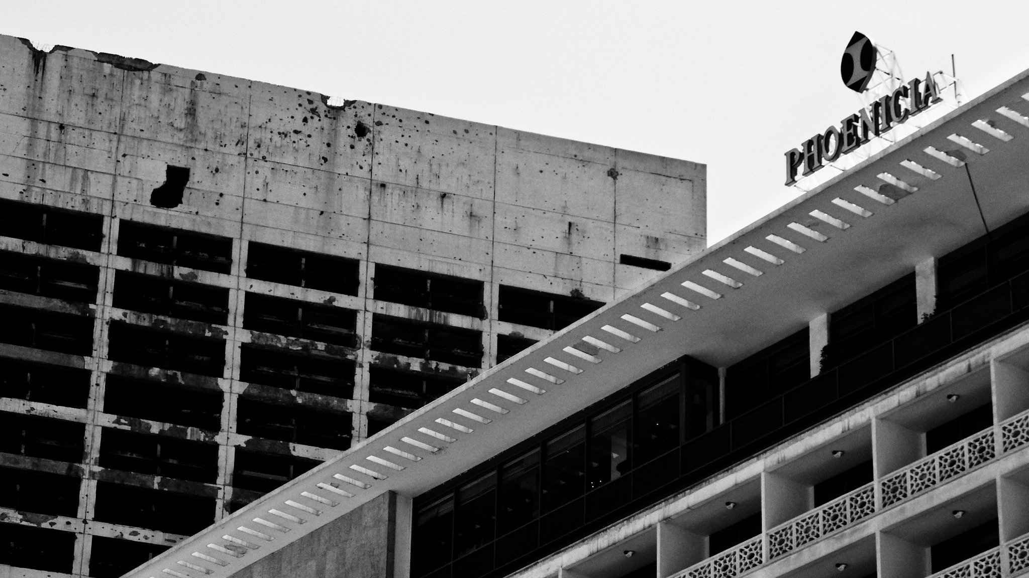 L'image montre deux bâtiments juxtaposés. À gauche, un bâtiment en béton brut et partiellement endommagé, avec des taches sombres visibles sur sa surface. À droite, un bâtiment plus moderne avec des lignes épurées et des balcons, arborant une façade blanche et des détails décoratifs. En haut à droite, le nom "Phoenix" est visible, probablement sur un panneau. L'ensemble de l'image est en noir et blanc, ce qui accentue le contraste entre les deux structures et leur état.