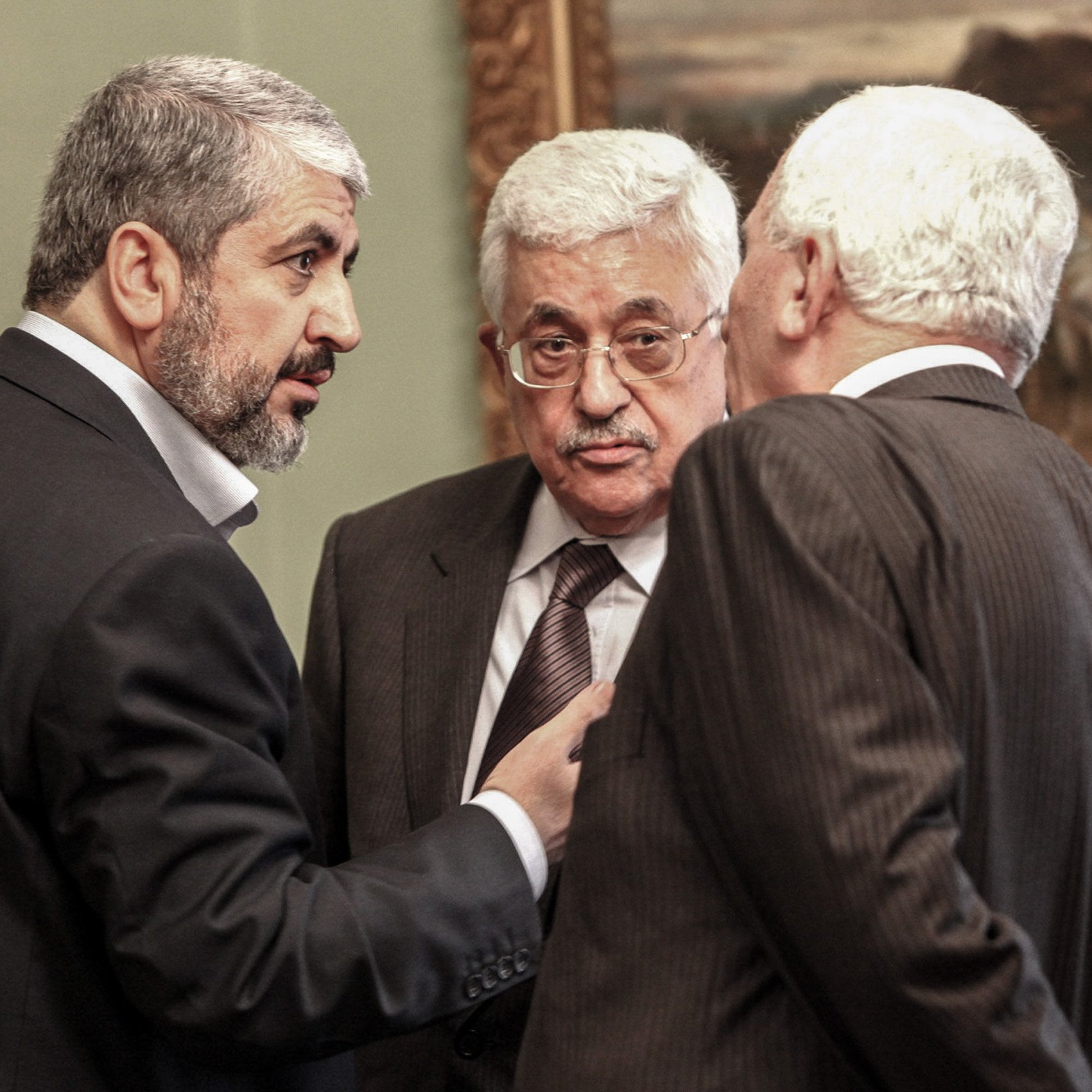 في الصورة، يظهر ثلاثة رجال يتبادلون الحديث في إطار رسمي. يرتدي الرجال بدلات رسمية، ويبدو عليهم التركيز أثناء المناقشة. خلفهم، يمكن رؤية لوحة فنية على الحائط تضيف لمسة ثقافية للمشهد. الجو العام يوحي بأن الحديث يجري حول موضوع مهم أو تفاوض سياسي.