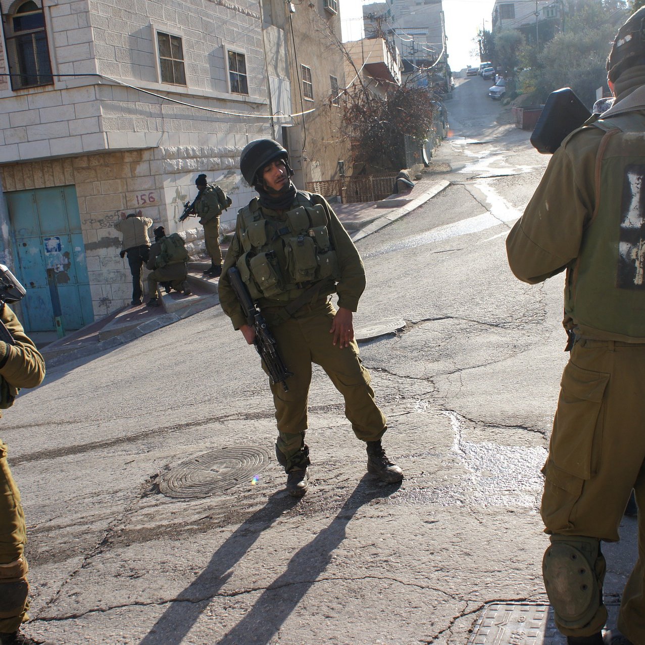 L'image montre des soldats armés en tenue militaire dans une rue apparemment urbaine. Deux d'entre eux sont en position de vigilance, tandis qu'un autre semble communiquer ou surveiller. L'environnement semble être un quartier situé dans une ville, avec des bâtiments en arrière-plan. L'atmosphère pourrait suggérer une situation de tension ou de surveillance.