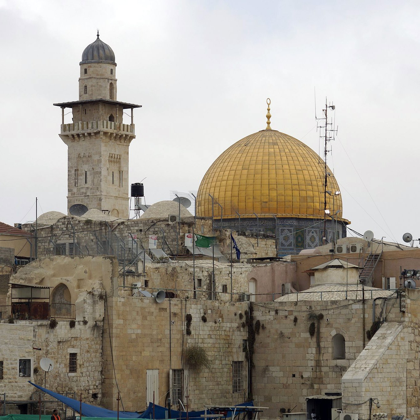 L'image montre une vue d'un paysage urbain avec des bâtiments en pierre. Au centre, on peut apercevoir un dôme doré, probablement celui du Rocher, qui est un site religieux important. À proximité, on remarque un minaret qui surplombe les structures anciennes. Le ciel est nuageux, ajoutant une atmosphère douce à la scène. Les toits sont parsemés de paraboles satellites, témoignant de la modernité mêlée à l'histoire de cette région.