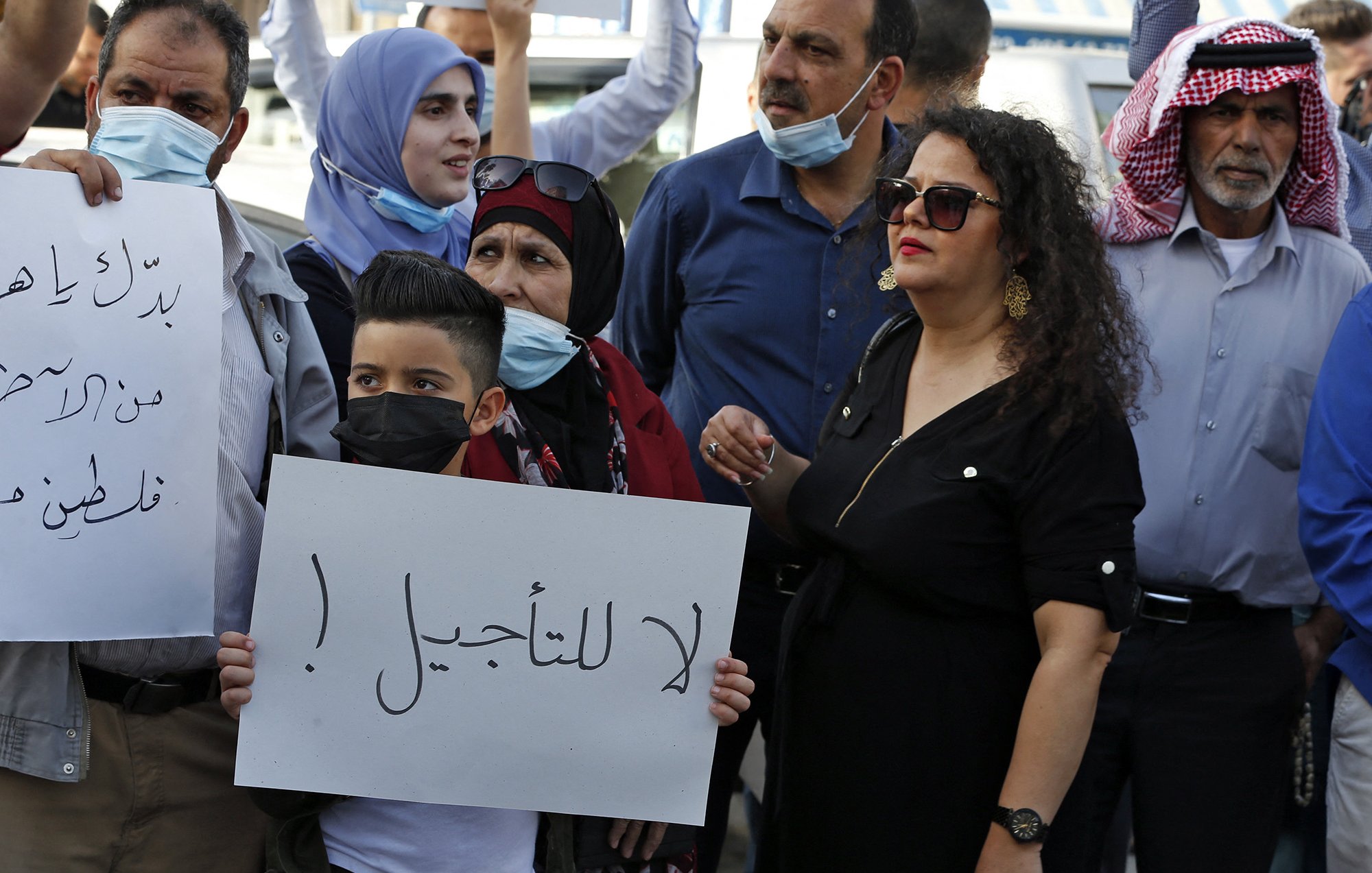 En la imagen se puede ver a un grupo de personas reunidas, algunas de las cuales sostienen carteles. La mayoría llevan mascarillas. Entre las personas, un niño sostiene un cartel que tiene un mensaje. Se observa una variedad de expresiones en sus rostros, reflejando una atmósfera de protesta o manifestación. La vestimenta de los participantes es variada, con algunas personas usando prendas tradicionales. El ambiente sugiere una situación de movilización social.