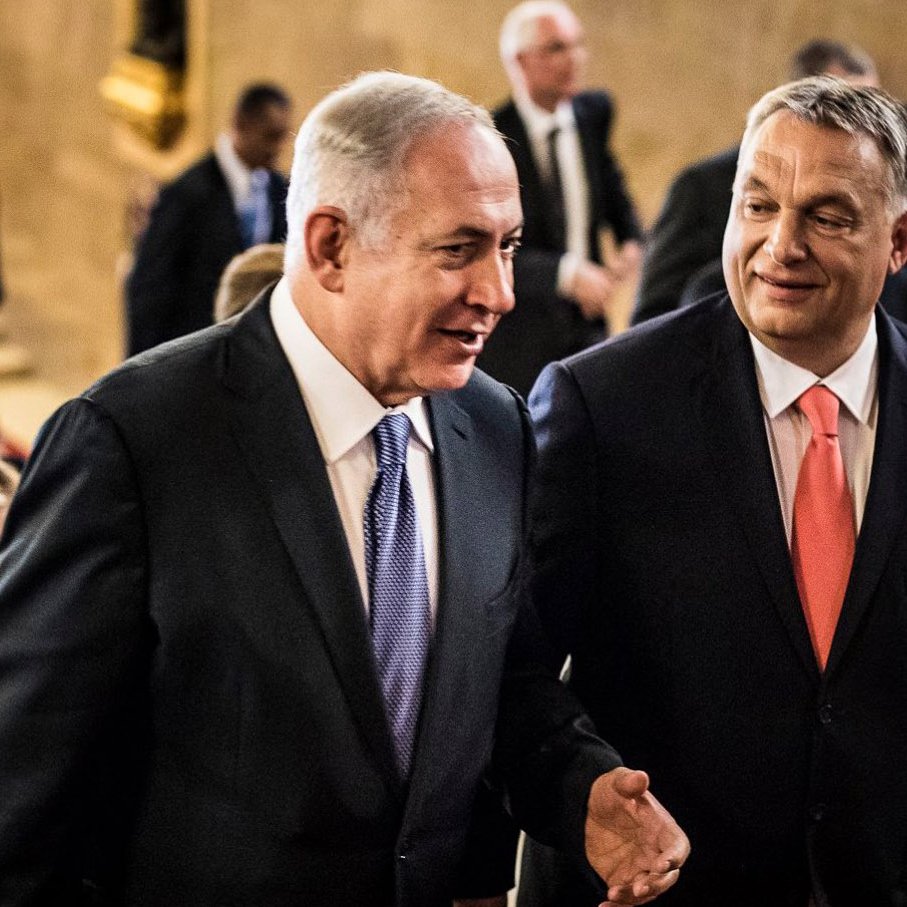 الصورة تُظهر رجلين يتحدثان معًا في ممر يبدو أنه في مبنى رسمي. يظهر أحدهما مرتديًا بدلة داكنة مع رابطة عنق زرقاء، بينما الآخر يرتدي بدلة فاتحة ورابطة عنق برتقالية. خلفهم، يظهر عدد من الأشخاص في بيئة احتفالية. اللغة الجسدية تعكس تفاعلاً ودياً بين الرجلين.