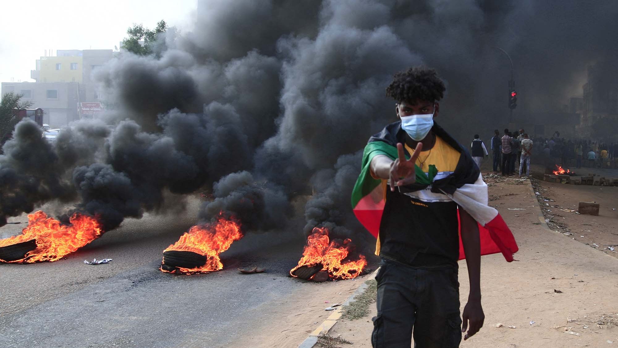 تصویر نشان‌دهنده یک صحنه اعتراضی است. در خیابان، چند لاستیک در حال سوختن هستند و دود سیاهی به هوا بلند شده است. در پس‌زمینه، گروهی از افراد دیده می‌شوند و در جلو، یک جوان با ماسک صورتی و لباسی خاص ایستاده که علامت پیروزی را با دستش نشان می‌دهد. جو کلی تصویر تنش‌آلود و حاکی از فعالیت‌های اعتراضی است.