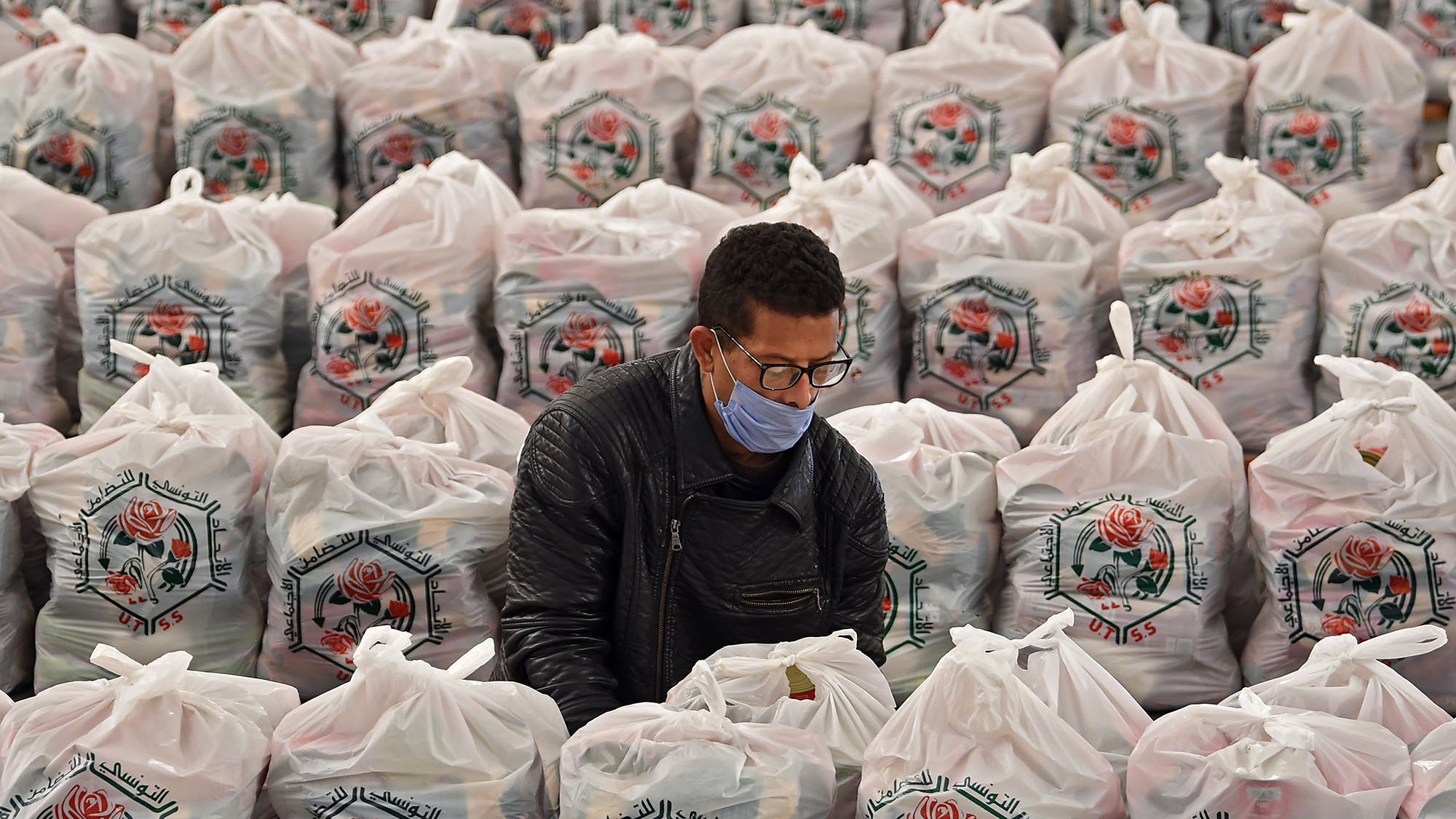L'image montre un homme en train de travailler au milieu d'un grand espace rempli de sacs en plastique. Chaque sac est soigneusement empilé, arborant un motif de rose et des inscriptions en arabe. L'homme porte un masque et semble concentré sur sa tâche, probablement en train de trier ou de préparer quelque chose à l'intérieur des sacs. L'environnement est bien organisé, et la scène évoque un sentiment d'efficacité dans un contexte d'entraide ou de distribution.
