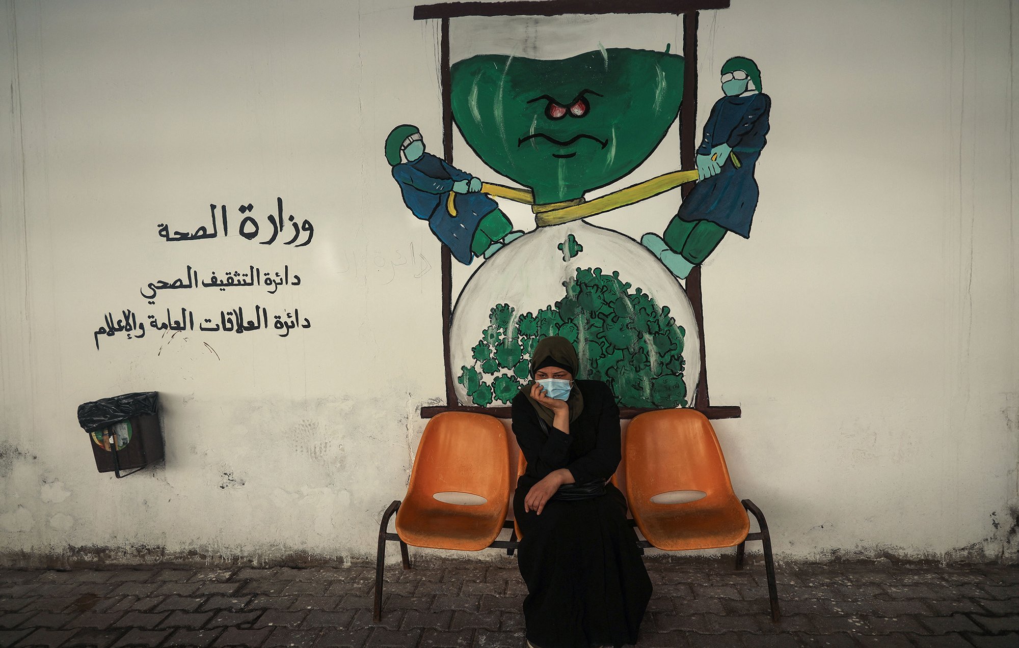 الصورة تظهر امرأة جالسة على كرسيين في منطقة تبدو وكأنها مركز صحي أو مستشفى. تحمل المرأة ملامح الجدية، وتضع كمامة على وجهها. خلفها يوجد جدار م painted يوضح رسماً يجسد فكرة وزارة الصحة، حيث يظهر شخصيتان ترتديان بزة وقاية، تسعيان لشد أو دفع شيء كبير يشبه الخضار أو الملفوف. الألوان المستخدمة في الجدار تعكس موضوع الصحة، مع تركيبات واضحة ورموز مثل القفازات والكمامات.