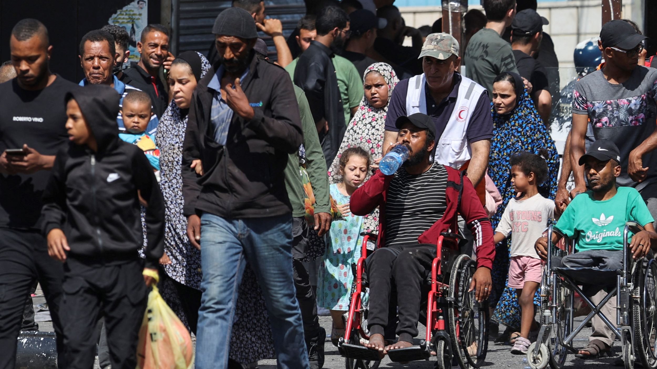 L'immagine mostra una scena affollata in un contesto urbano. Ci sono diverse persone che camminano insieme, tra cui adulti, bambini e alcune persone in carrozzina. Molte persone indossano abiti tradizionali o casual. C'è un'atmosfera vivace, ma al contempo seria, e i volti esprimono diverse emozioni. In primo piano, una persona in carrozzina tiene in mano una bottiglia d'acqua. Il contesto potrebbe suggerire una comunità che si riunisce, ma le condizioni dell'ambiente attorno sono visibili, con segni di spostamenti e attività quotidiane.