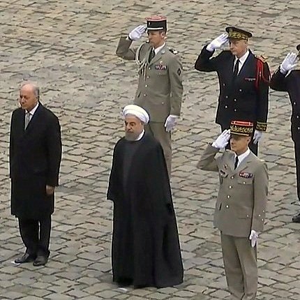 الصورة تظهر مجموعة من الأشخاص في موقف رسمي. يوجد شخصان في المقدمة، أحدهما في بدلة رسمية والآخر يرتدي الزي التقليدي. خلفهم يوجد عدد من الضباط العسكريين بزيهم الرسمي، يقومون بتحية عسكرية. الأرضية مكونة من حجارة مرصوفة. الجو يبدو رسميًا وجادًا، مما يشير إلى حدث ذو أهمية سياسية أو عسكرية.