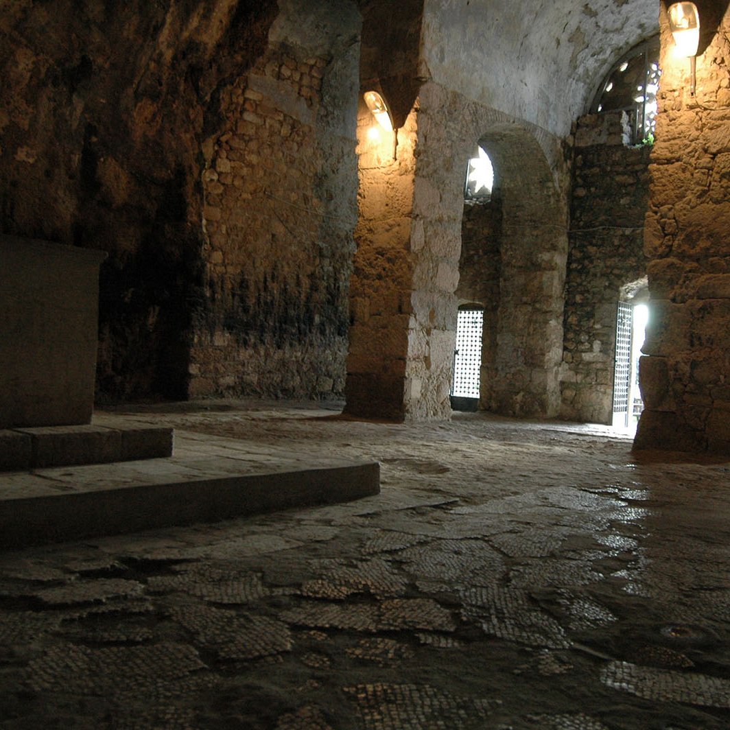 L'image montre un intérieur de cave ou de grotte, avec des murs en pierre apparente. On peut voir des arches qui soutiennent le plafond, et la lumière filtre par des fenêtres grillagées, créant une ambiance sombre mais fascinante. Le sol est en pierres irrégulières, et il semble y avoir un espace dégagé au centre. L'environnement dégage une atmosphère ancienne, presque mystique.