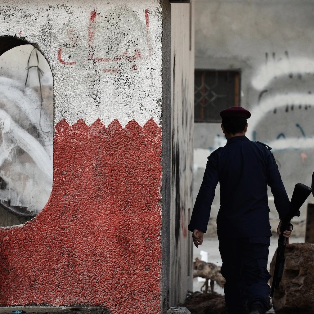 L'image montre deux policiers marchant le long d'un mur. Ce mur présente une décoration en rouge et blanc, avec une forme ovale découpée. En arrière-plan, plusieurs murs sont couverts de graffitis et apparaissent en mauvais état, témoignant d'une ambiance de tension ou de désolation. Les policiers, munis de casques et d'armement, semblent patrouiller dans une zone qui pourrait être marquée par des troubles ou des désordres.